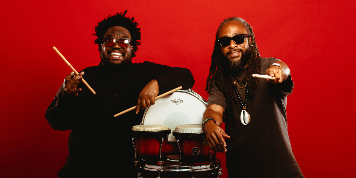 Modupe Onilu and Rhys Thompson of Boom Boom Room leaning on percussion drums against a solid red backdrop.