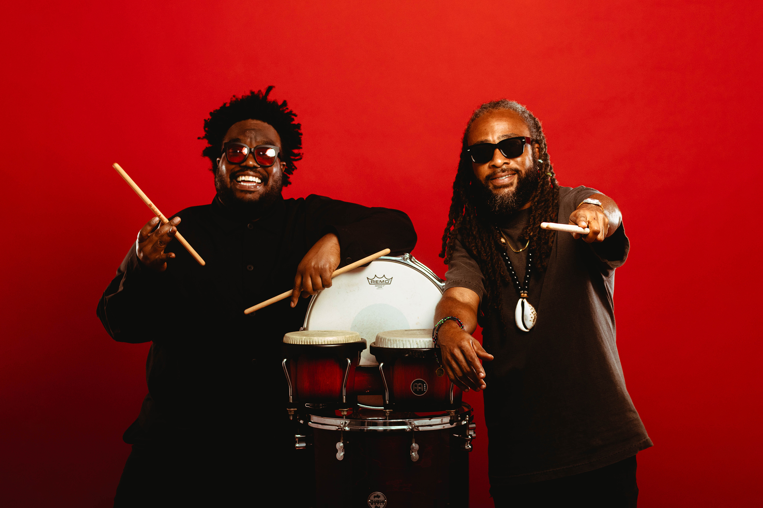 Modupe Onilu and Rhys Thompson of Boom Boom Room leaning on percussion drums against a solid red backdrop.